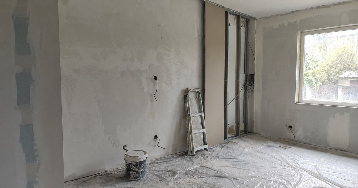 Pièce de maison en rénovation à l'arrêt, murs enduits inachevés, escabeau replié et bâche de protection au sol